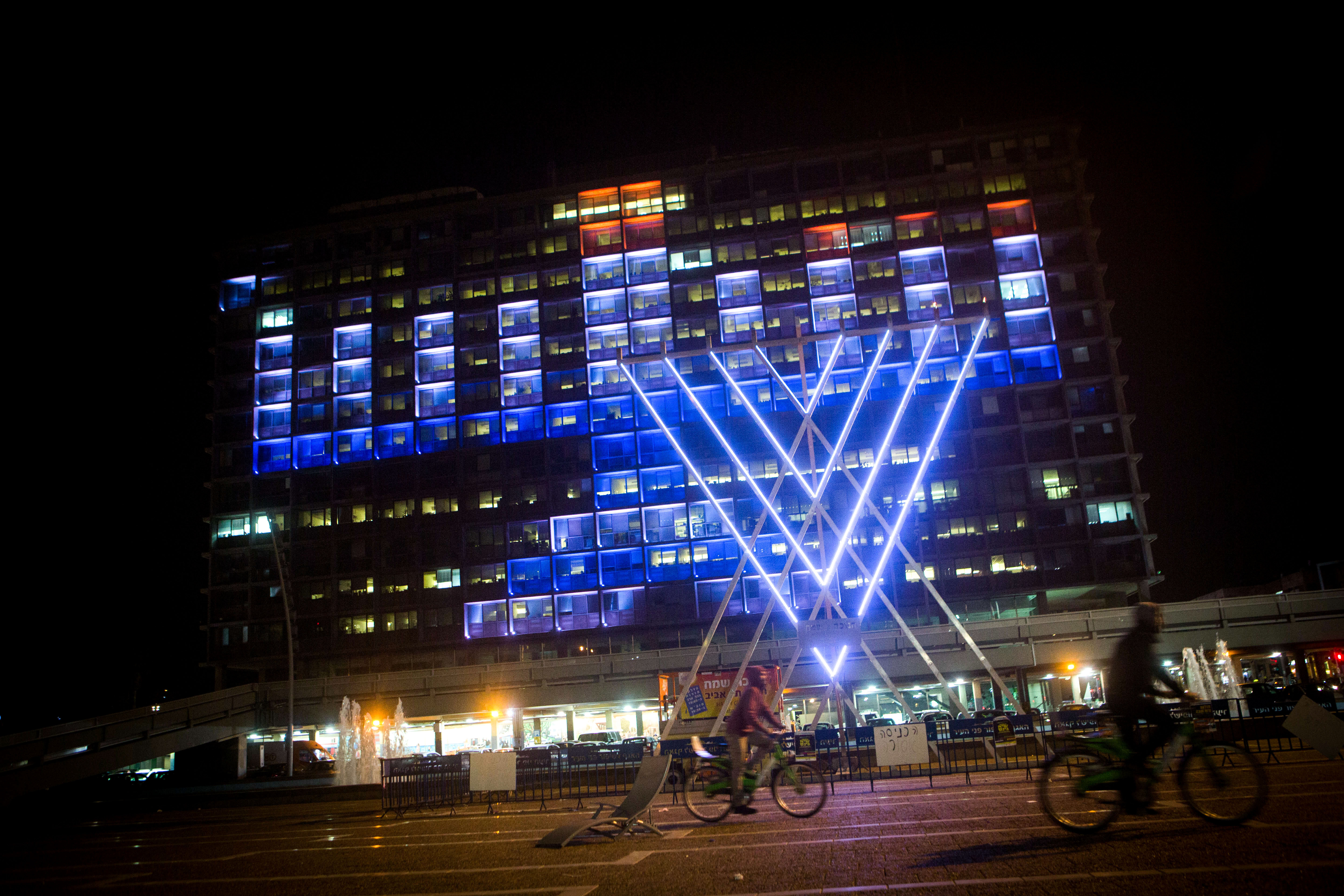 Israelis ride their bicycles past a lit up 'chanukia' displayed on the Tel Aviv municipality building in Tel Aviv on December 8, 2015. Hanukkah, also known as the Festival of Lights, is an eight-day Jewish holiday commemorating the rededication of the Holy Temple. The festival is observed by the kindling of the lights of a 'hanuckia'- a nine-branched candelabrum, with one additional light being lit on each night of the holiday.