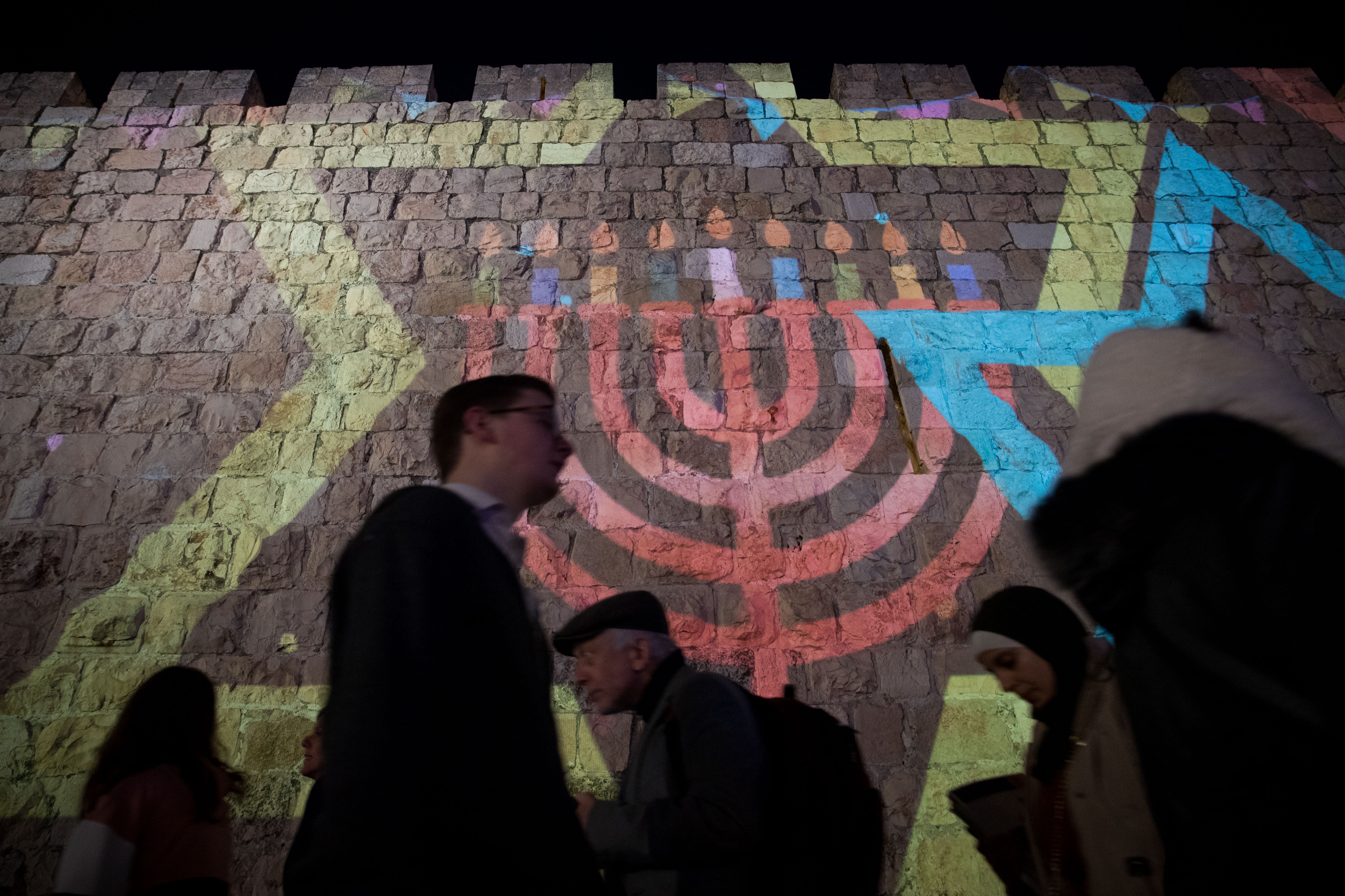 Pepole walk past the walls of Jerusalem's Old City illuminated with projection showing Menorah or Hanukkah lamp during the Jewish holiday of Hanukkah on December 24, 2019.