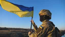 Soldier in military uniform stands to the peak of hill waving flag of Ukraine