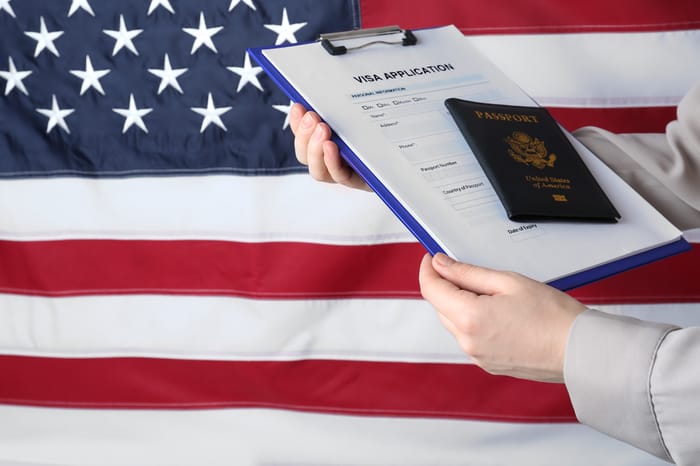 Woman holding visa application form and passport against American flag, closeup.