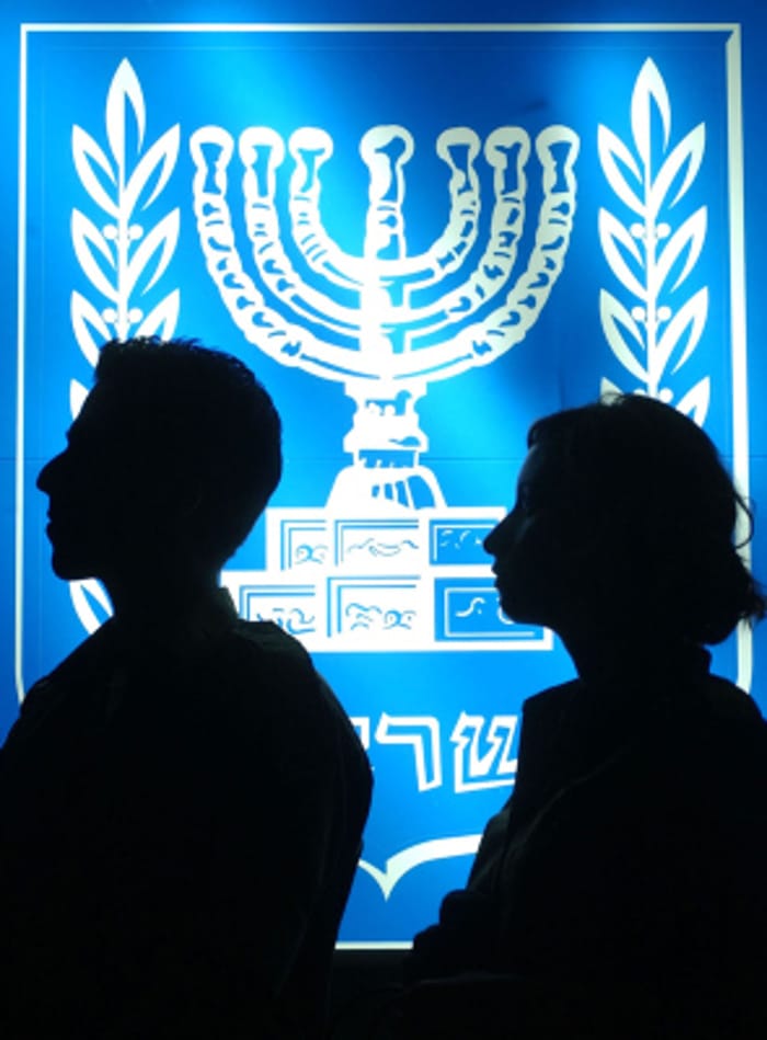 A male and a female soldier stand in front of a lit up emblem of the state of Israel.