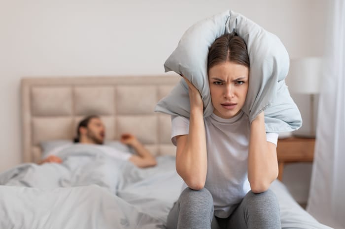 Exasperated woman sitting in bed holding pillow over her ears to block out the sound of man snoring loudly