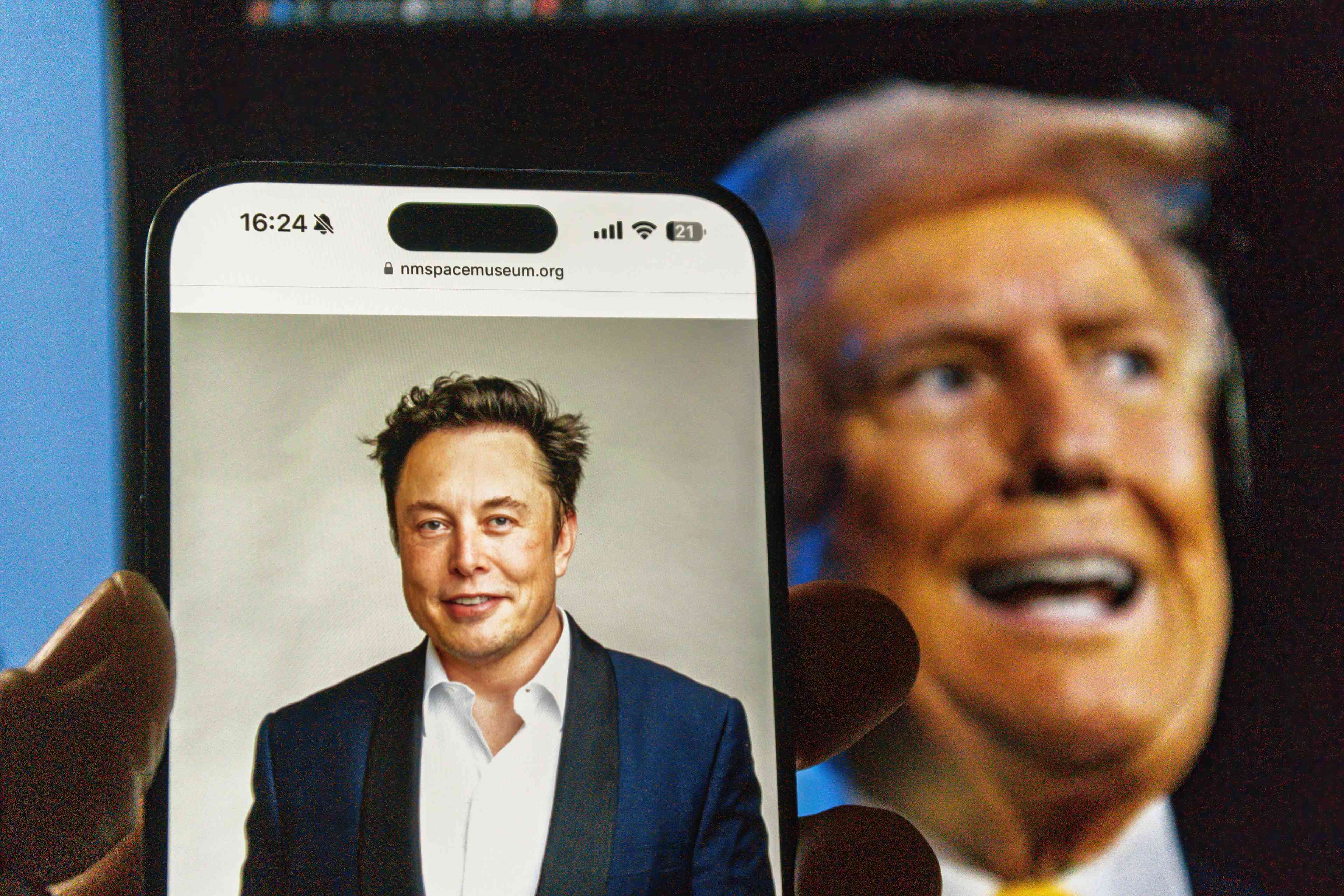 A smartphone displays a portrait of Elon Musk from the NM Space Museum website, with a background image of Donald Trump background