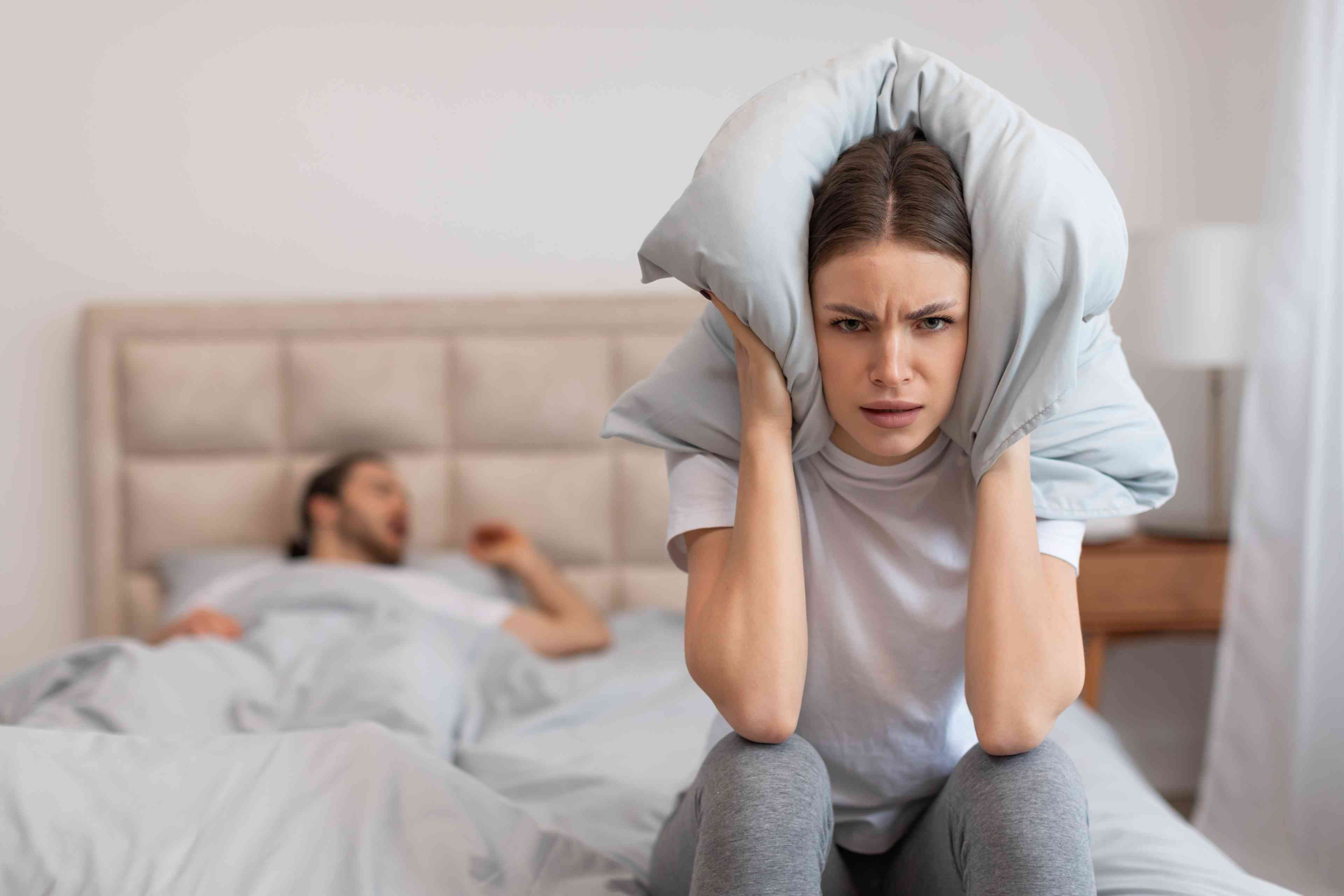 Exasperated woman sitting in bed holding pillow over her ears to block out the sound of man snoring loudly background