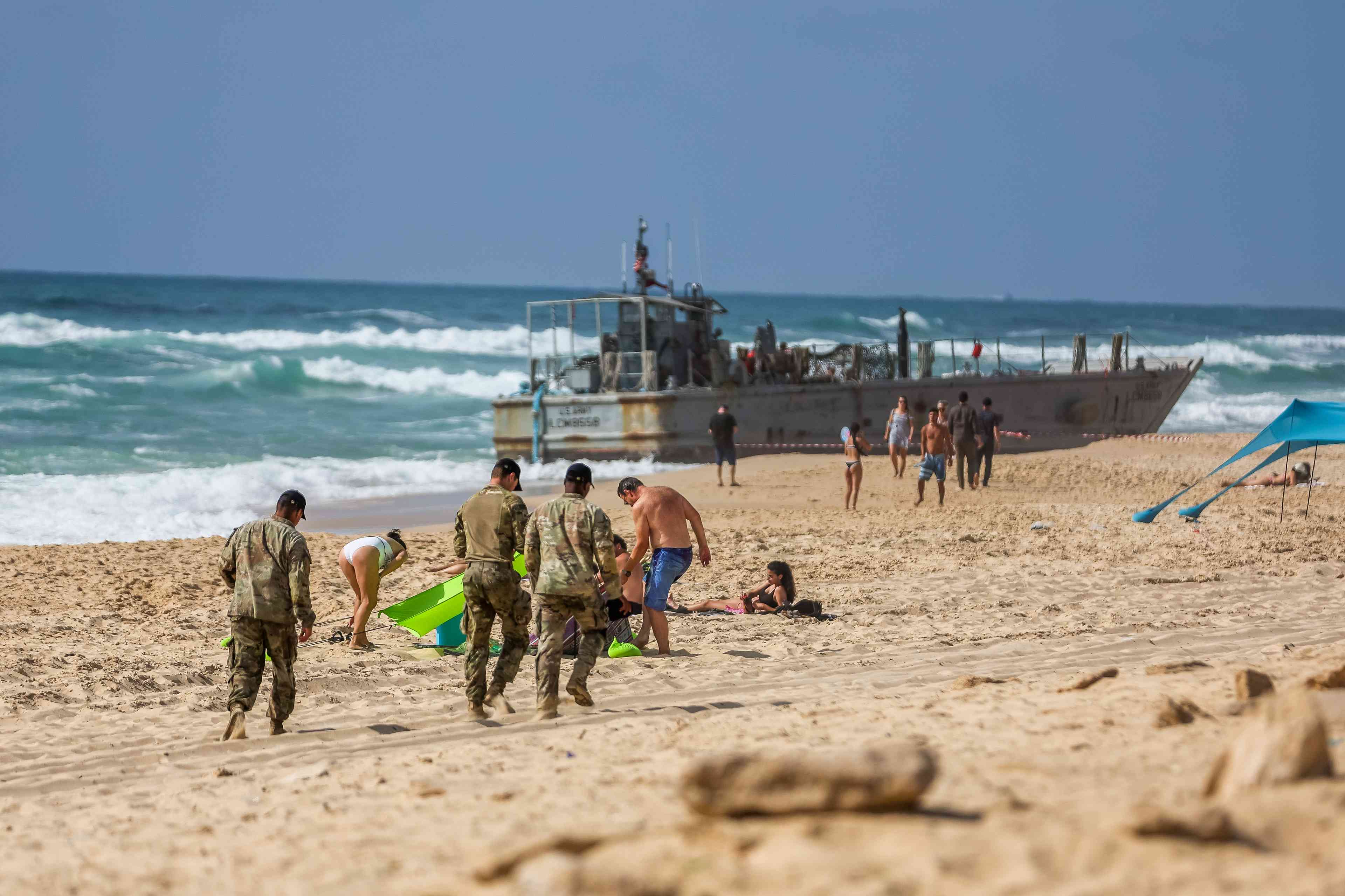 US navy soldier and Israelis near a US Army vessel that washed up on the beach in the southern Israeli city in Ashdod, May 25, 2024 background