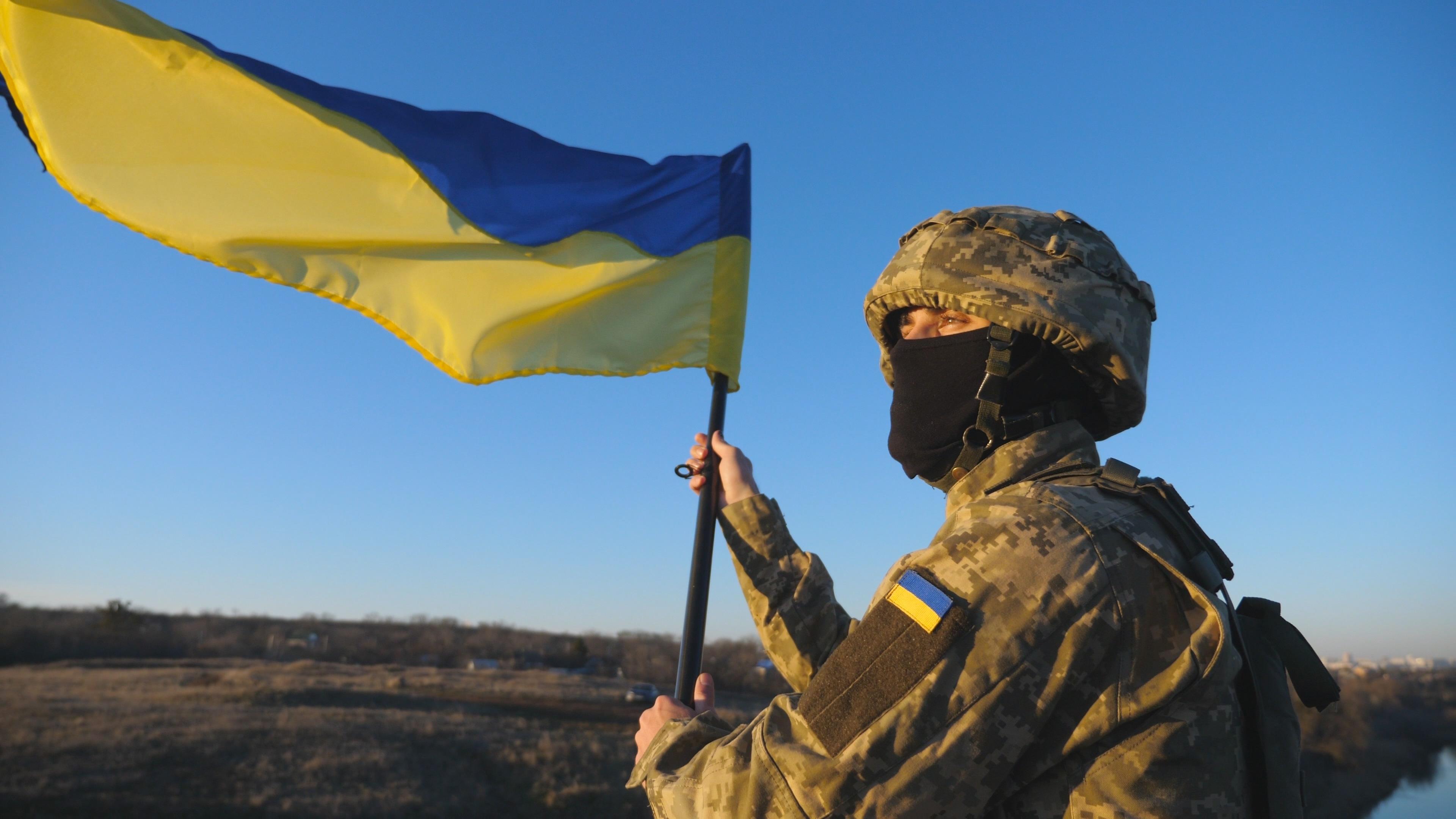 Soldier in military uniform stands to the peak of hill waving flag of Ukraine