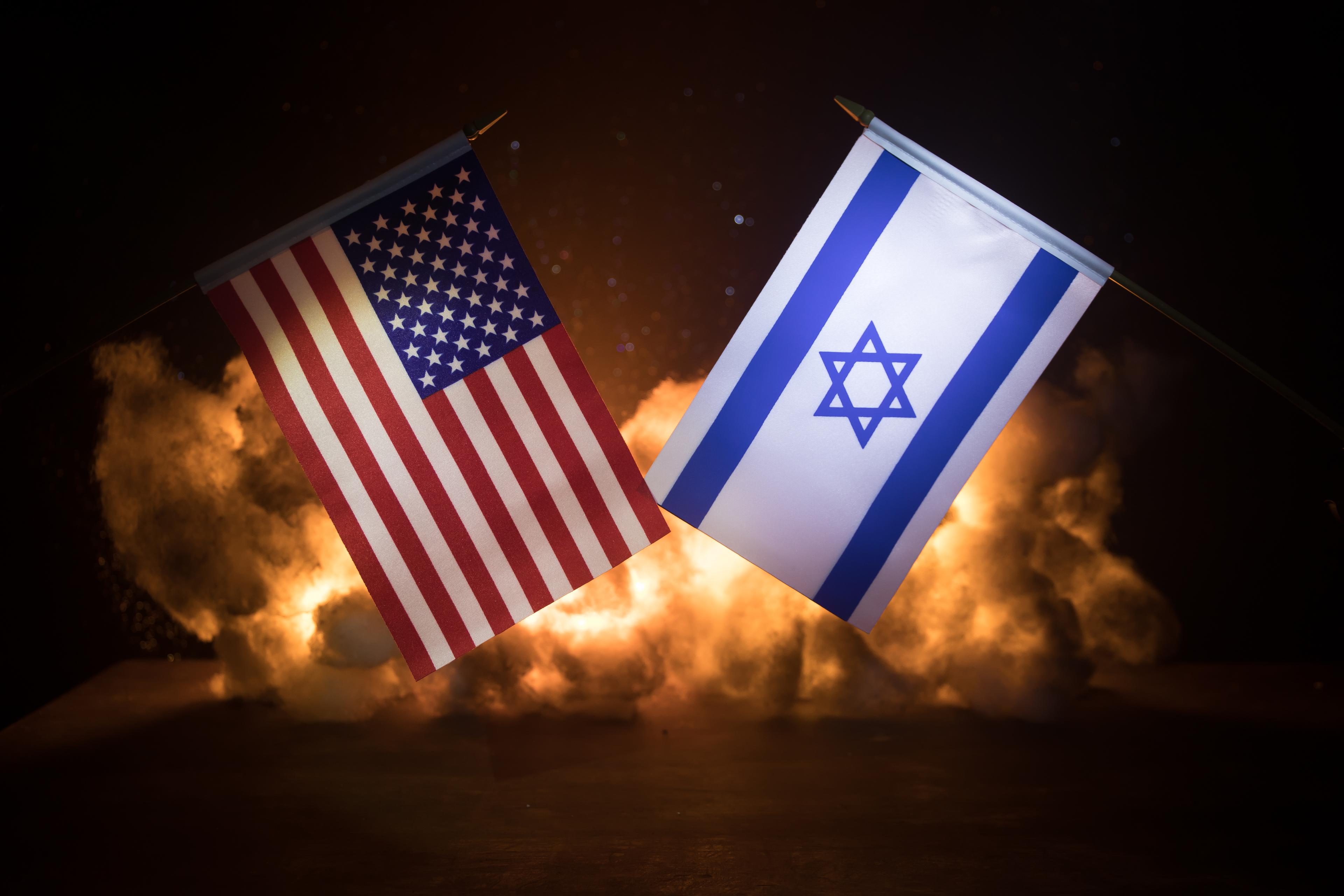 Israel and USA small flags on burning dark background