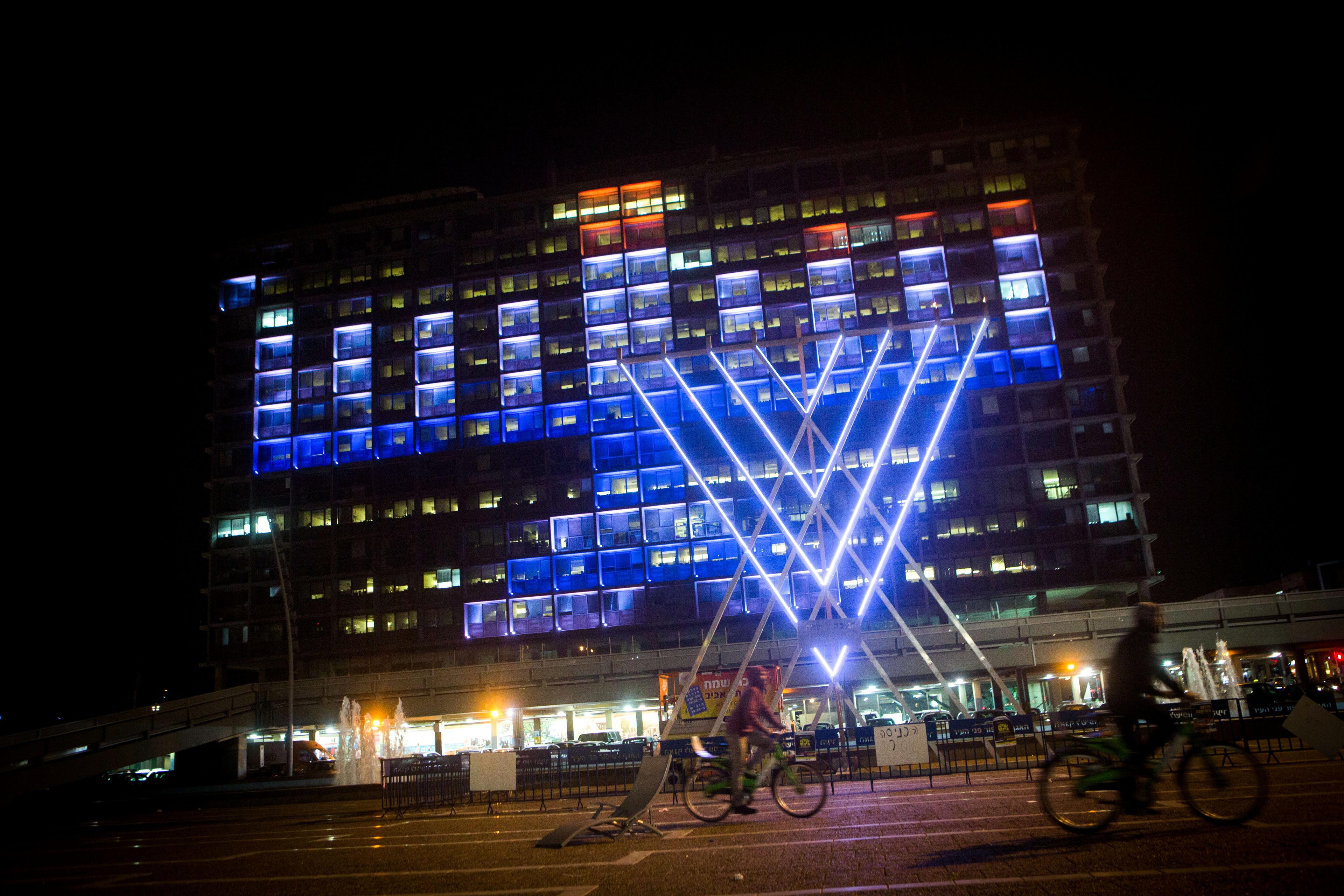 Israelis ride their bicycles past a lit up 'chanukia' displayed on the Tel Aviv municipality building in Tel Aviv on December 8, 2015. Hanukkah, also known as the Festival of Lights, is an eight-day Jewish holiday commemorating the rededication of the Holy Temple. The festival is observed by the kindling of the lights of a 'hanuckia'- a nine-branched candelabrum, with one additional light being lit on each night of the holiday.